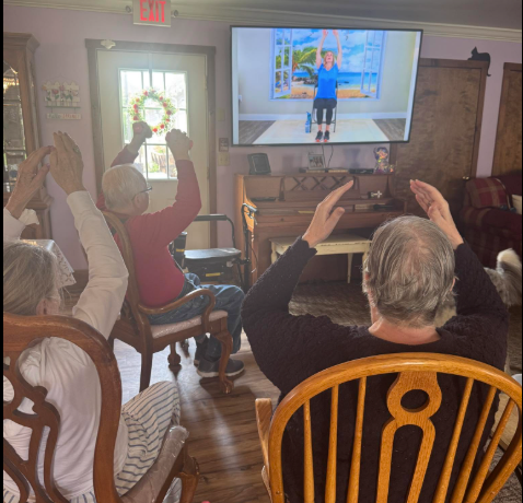 Residents seated in the living room with arms raised, following along with a chair exercise class on the television at Lavender Fields