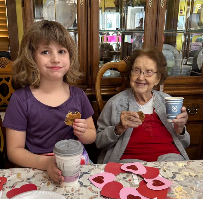 A young girl and a resident sharing cookies and hot drinks at the dining room table, Valentine hearts spread between them